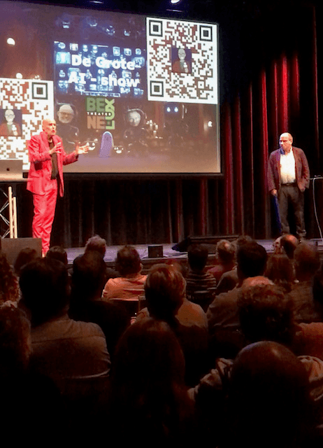 Two men presenting on stage with a large screen displaying QR codes and audience watching.